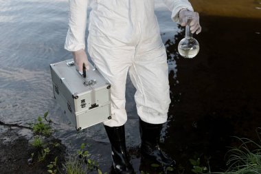 partial view of water inspector in protective costume holding inspection kit and flask at river clipart