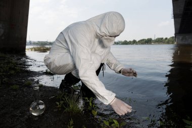 water inspector in protective costume and respirator taking water sample at river clipart