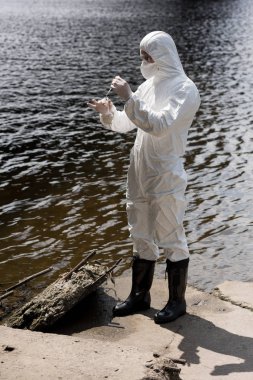 full length view of water inspector in protective costume, latex gloves and respirator taking water sample at river clipart
