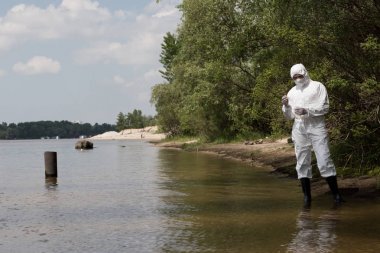 full length view of water inspector in protective costume, latex gloves and respirator holding flask with water sample at river clipart
