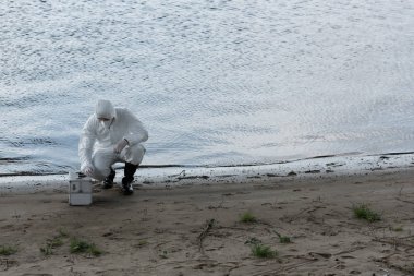 water inspector in protective costume and respirator with inspection kit sitting at river coast clipart