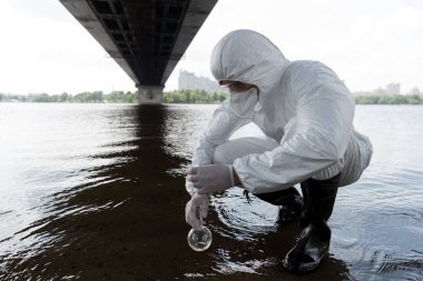 koruyucu kostüm içinde su denetçisi şişe tutan ve nehirden su örneği alma