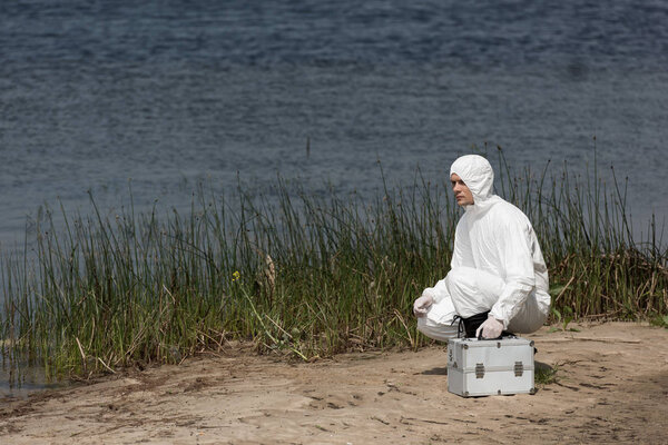 water inspector in protective costume with inspection kit sitting on river coast