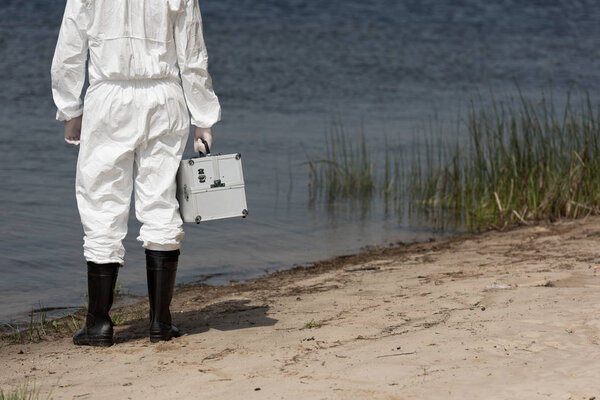 cropped view of water inspector in protective suit holding inspection kit on river coast