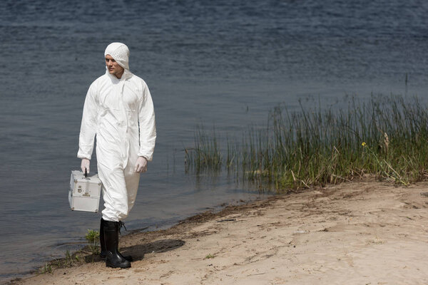 full length view of water inspector in protective suit holding inspection kit and standing on river coast