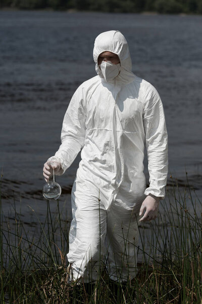water inspector in protective costume, respirator and goggles holding flask with water sample
