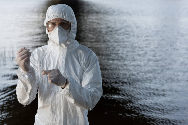 water inspector in protective costume, goggles and respirator taking water sample at river