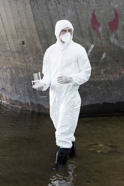 full length view of water inspector in protective costume and respirator holding test tubes and flask at river