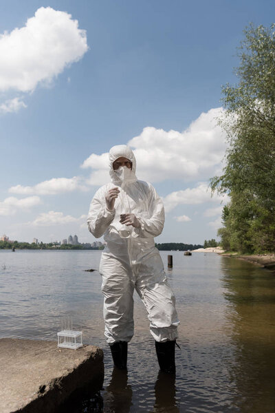 full length view of water inspector in protective costume, latex gloves and respirator holding flask with water sample at river