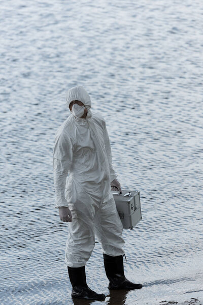 full length view of water inspector in protective costume holding inspection kit at river