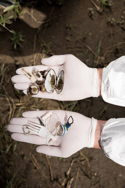 cropped view of ecologist in latex gloves holding seashells and plastic garbage