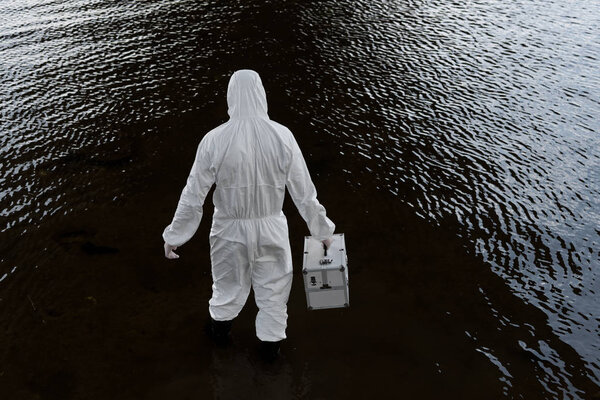 back view of water inspector in protective costume holding inspection kit in river