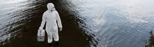 panoramic shot of water inspector in protective costume and respirator holding inspection kit at river