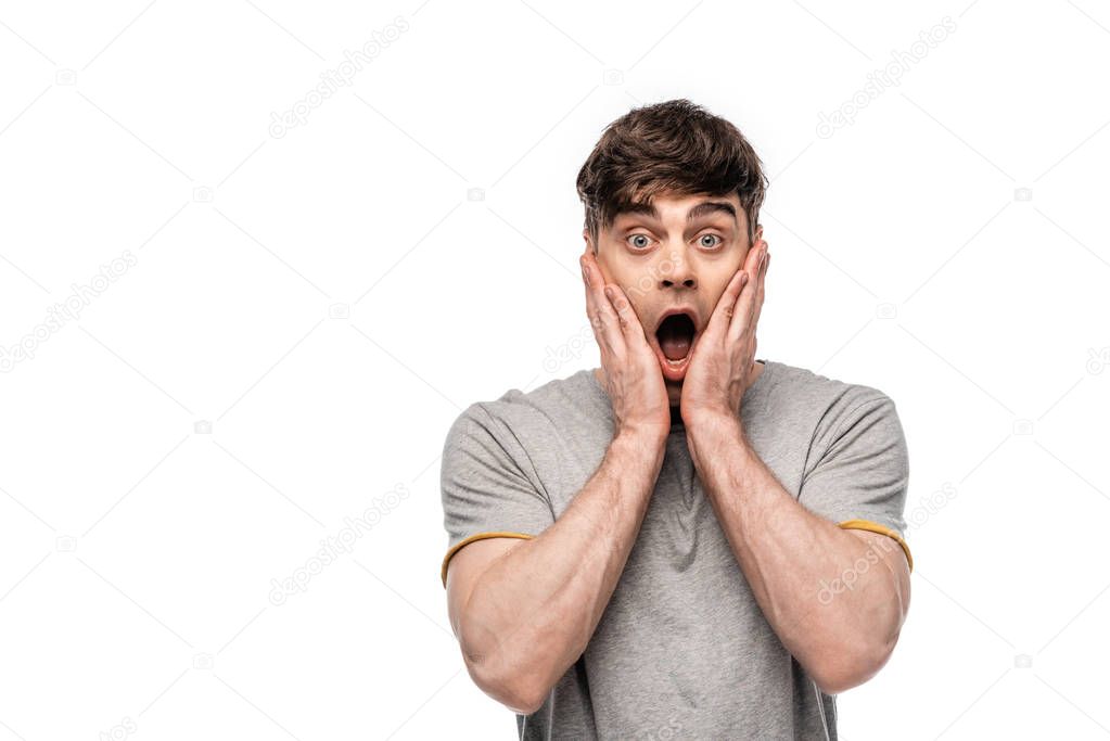 shocked young man looking at camera while holding hands near face isolated on white