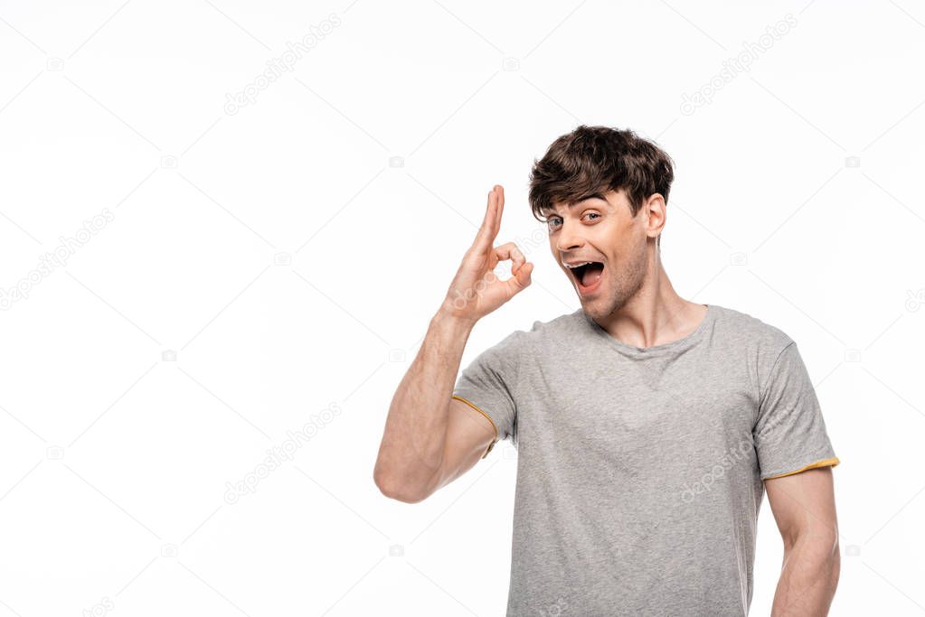 cheerful young man showing ok gesture while smiling at camera isolated on white