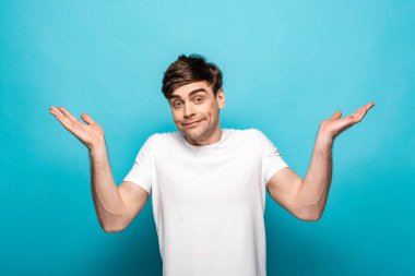 disappointed young man looking at camera and showing shrug gesture on blue background clipart