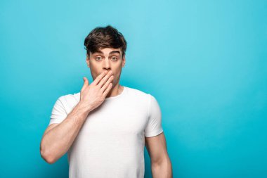 shocked young man covering mouth with hand while looking at camera on blue background clipart