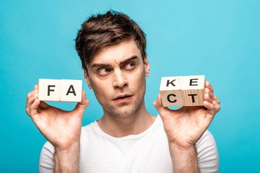 thoughtful young man looking away while holding wooden cubes with fake fact lettering isolated on blue