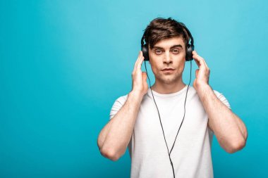 pensive young man listening music in headphones and looking at camera on blue background clipart