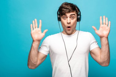 discouraged young man showing give up gesture while listening music in headphones on blue background clipart