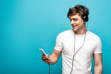 handsome young man smiling while listening music with headphones and smartphone on blue background clipart