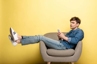 handsome young man sitting in armchair and using smartphone on yellow background clipart