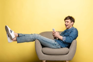neşeli genç adam koltukta oturan ve sarı arka plan üzerinde akıllı telefon kullanarak