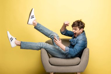 excited young man showing yes gesture while sitting in armchair and using smartphone on yellow background clipart