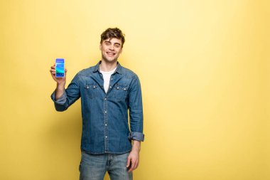 handsome smiling man in denim clothes holding smartphone with booking app on yellow clipart