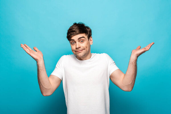 disappointed young man looking at camera and showing shrug gesture on blue background