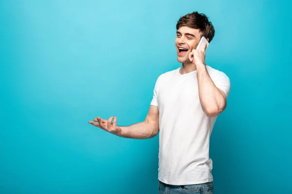 Jovem Alegre Gesticulando Enquanto Fala Smartphone Olhando Para Fundo Azul — Fotografia de Stock