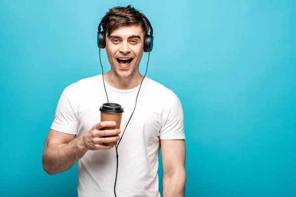 快乐的年轻人在耳机里听音乐 同时在蓝色背景上拿着一次性杯子 — 图库照片