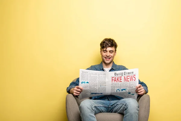 アームチェアに座って 黄色の背景に偽のニュースで新聞を読んで笑顔の若者 — ストック写真