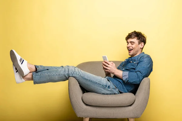 开朗的年轻人坐在扶手椅上 用黄色背景的智能手机 — 图库照片