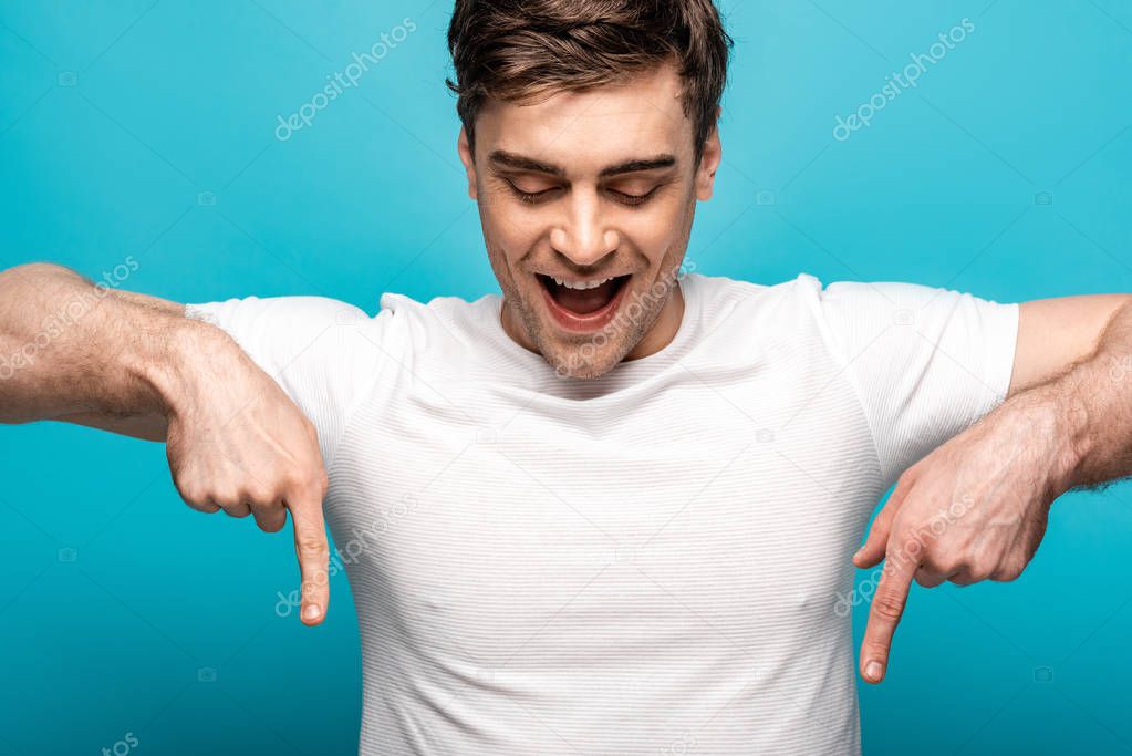 cheeful young man pointing down with fingers on blue background