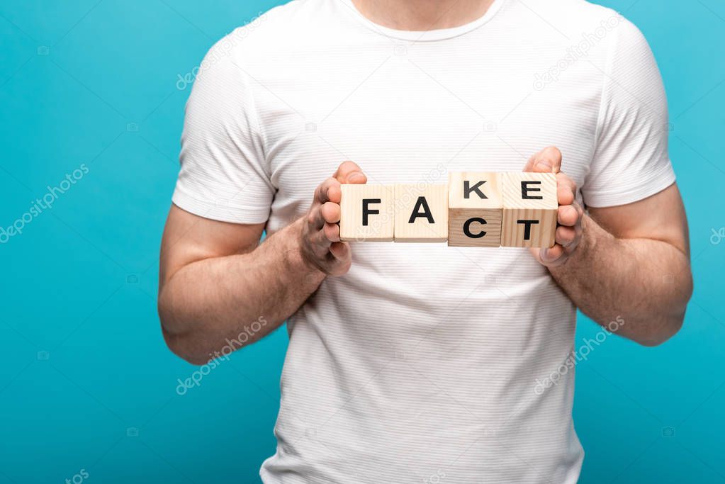 partial view of man in white t-shirt holding wooden cubes with fake fact lettering on blue background 