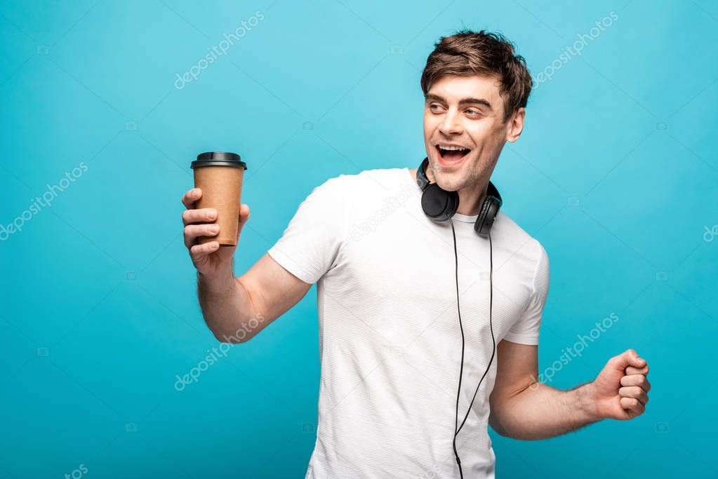 cheerful man with headphones looking away while holding paper cup on blue background