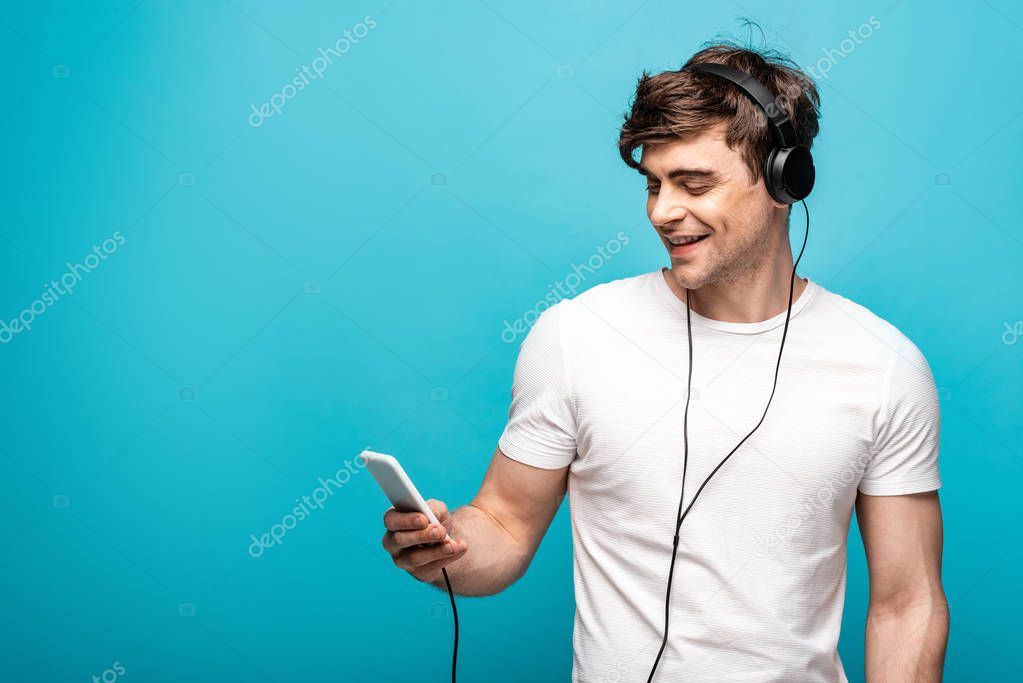 handsome young man smiling while listening music with headphones and smartphone on blue background