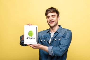 Kiev, Ukrayna - 16 Mayıs 2019: android uygulaması ile dijital tablet gösteren kot pantolon giysiler yakışıklı neşeli adam, sarı izole