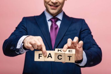 cropped view of businessman holding wooden cubes with fake fact lettering on pink background clipart