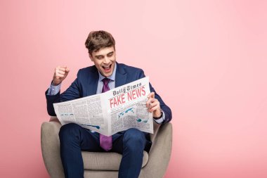 neşeli işadamı koltukta otururken ve pembe arka plan üzerinde sahte haber ile gazete okurken kazanan jest gösteren