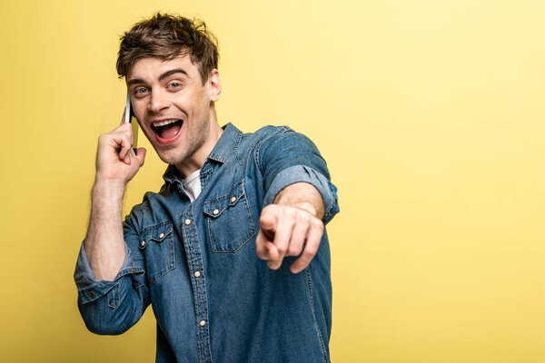 excited young man pointing with finger at camera while talking on smartphone on yellow background