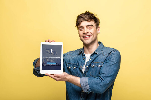 KYIV, UKRAINE - MAY 16, 2019: handsome smiling man in jeans clothes showing digital tablet with tumblr app, isolated on yellow