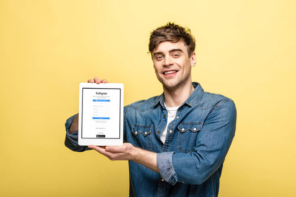 KYIV, UKRAINE - MAY 16, 2019: handsome smiling man in jeans clothes showing digital tablet with instagram app, isolated on yellow