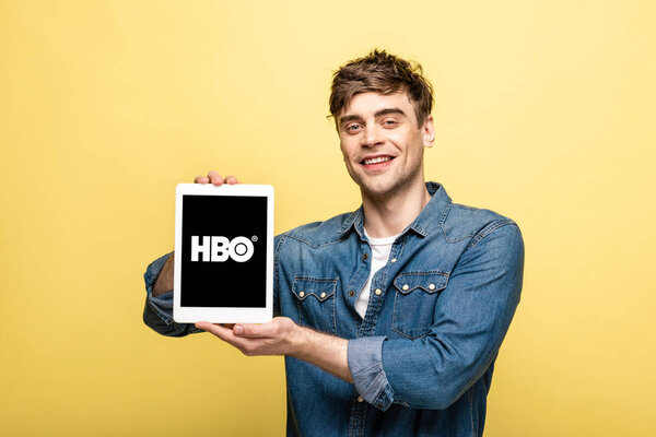 KYIV, UKRAINE - MAY 16, 2019: handsome cheerful man in jeans clothes showing digital tablet with HBO app, isolated on yellow