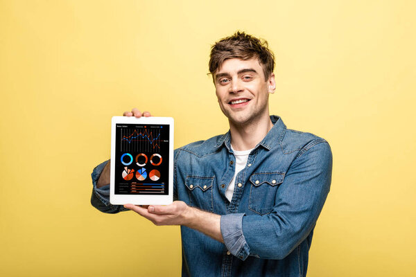 handsome smiling man in denim clothes showing digital tablet with infographic, isolated on yellow