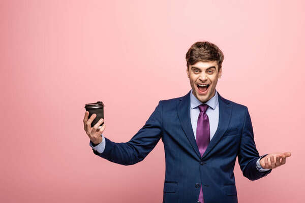 excited businessman holding coffee to go while laughing at camera on pink background