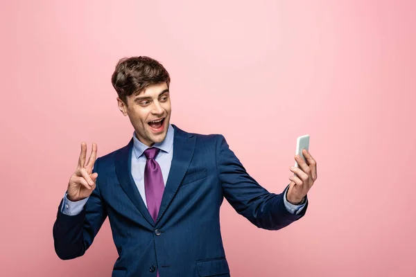 ピンクの背景にスマートフォンを使用しながら勝利のジェスチャーを示す陽気なビジネスマン — ストック写真