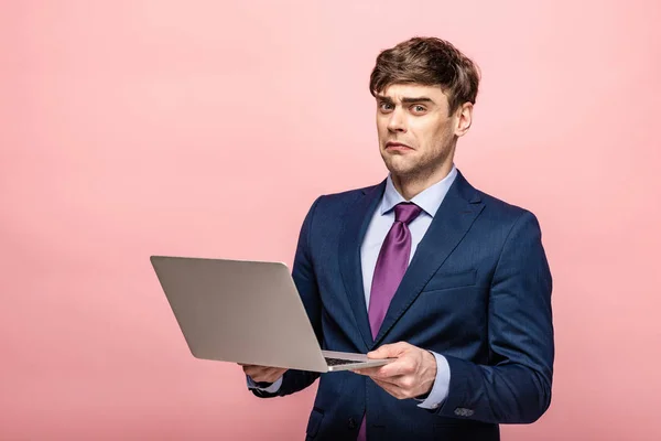 disappointed businessman looking at camera while holding laptop on pink background