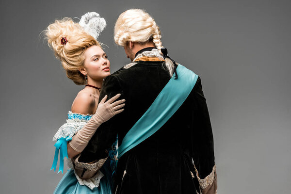 привлекательная викторианка в синем платье обнимающая мужчину в парике на сером
